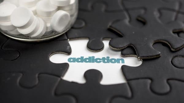 Pills spilling from a bottle onto a puzzle with a piece missing that spells out 'addiction.'