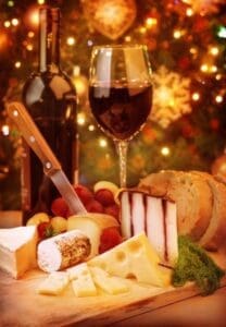 A festive holiday table featuring a bottle of wine, a filled glass, assorted cheeses, bread, and grapes with twinkling lights in the background.