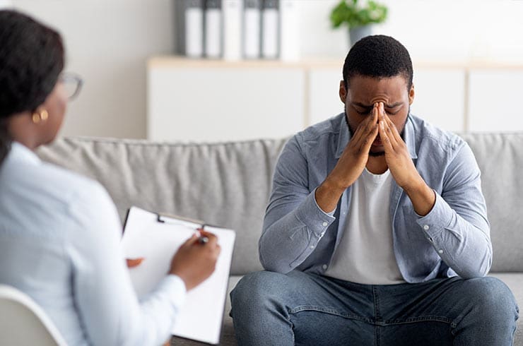 man with PTSD sits on a couch clutching his face while talking to a female therapist