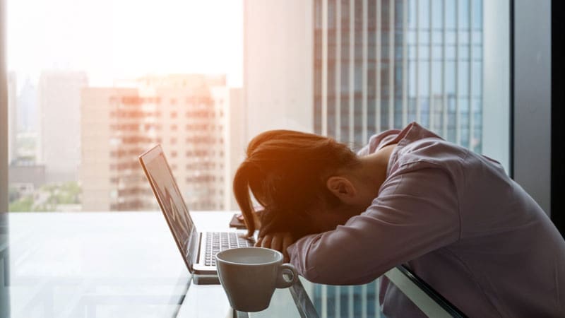 A fatigued person resting their head on a desk beside a laptop with a cup of coffee, suggestive of exhaustion or sleep issues.