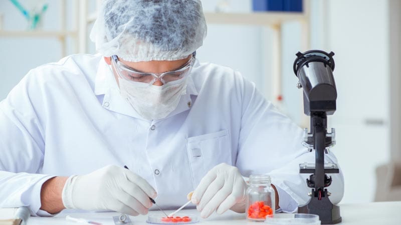 A lab technician in protective clothing analyzes samples next to a microscope and a jar of pills.