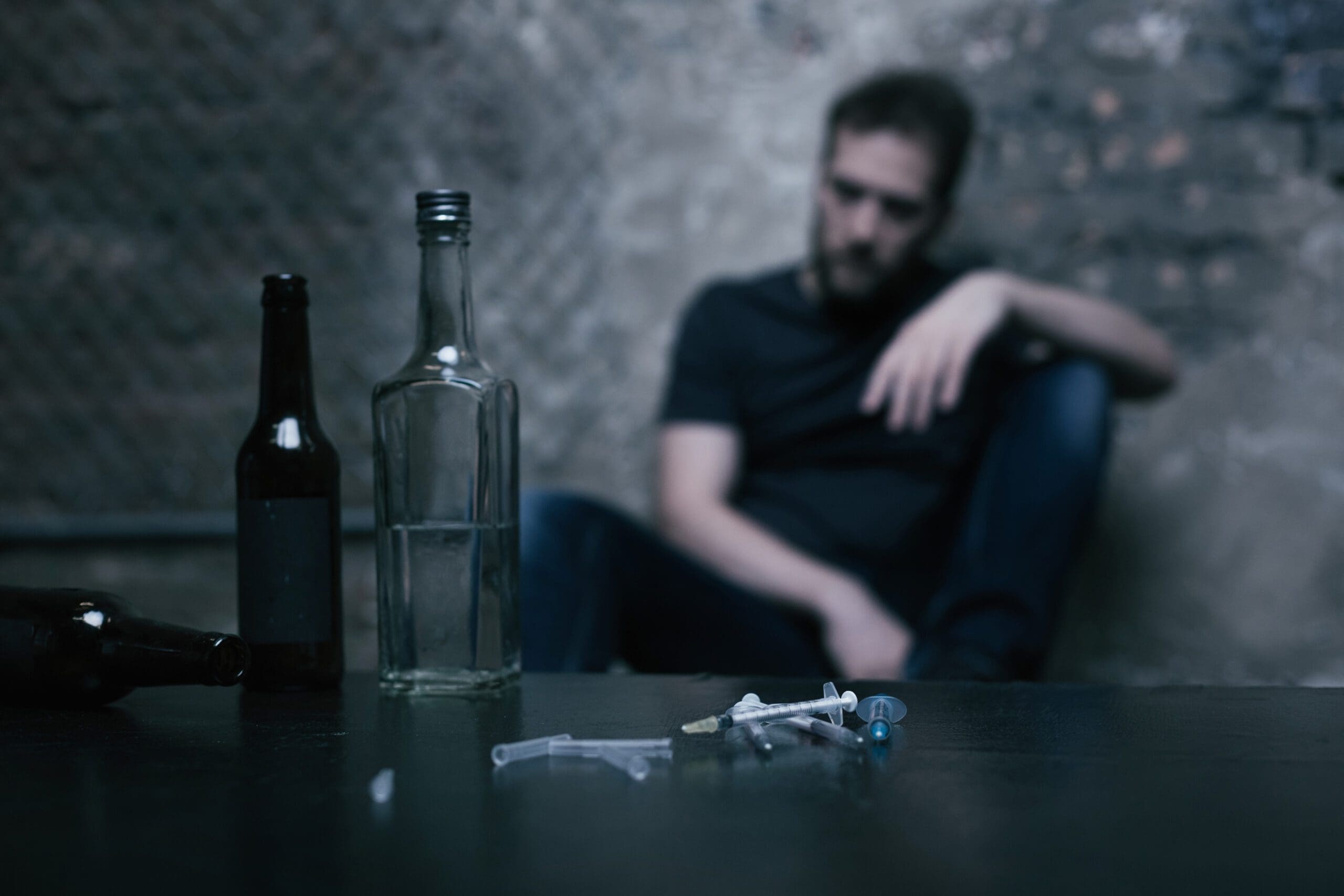 A man appears sitting in the background, looking contemplative, with empty bottles and syringes in the foreground.