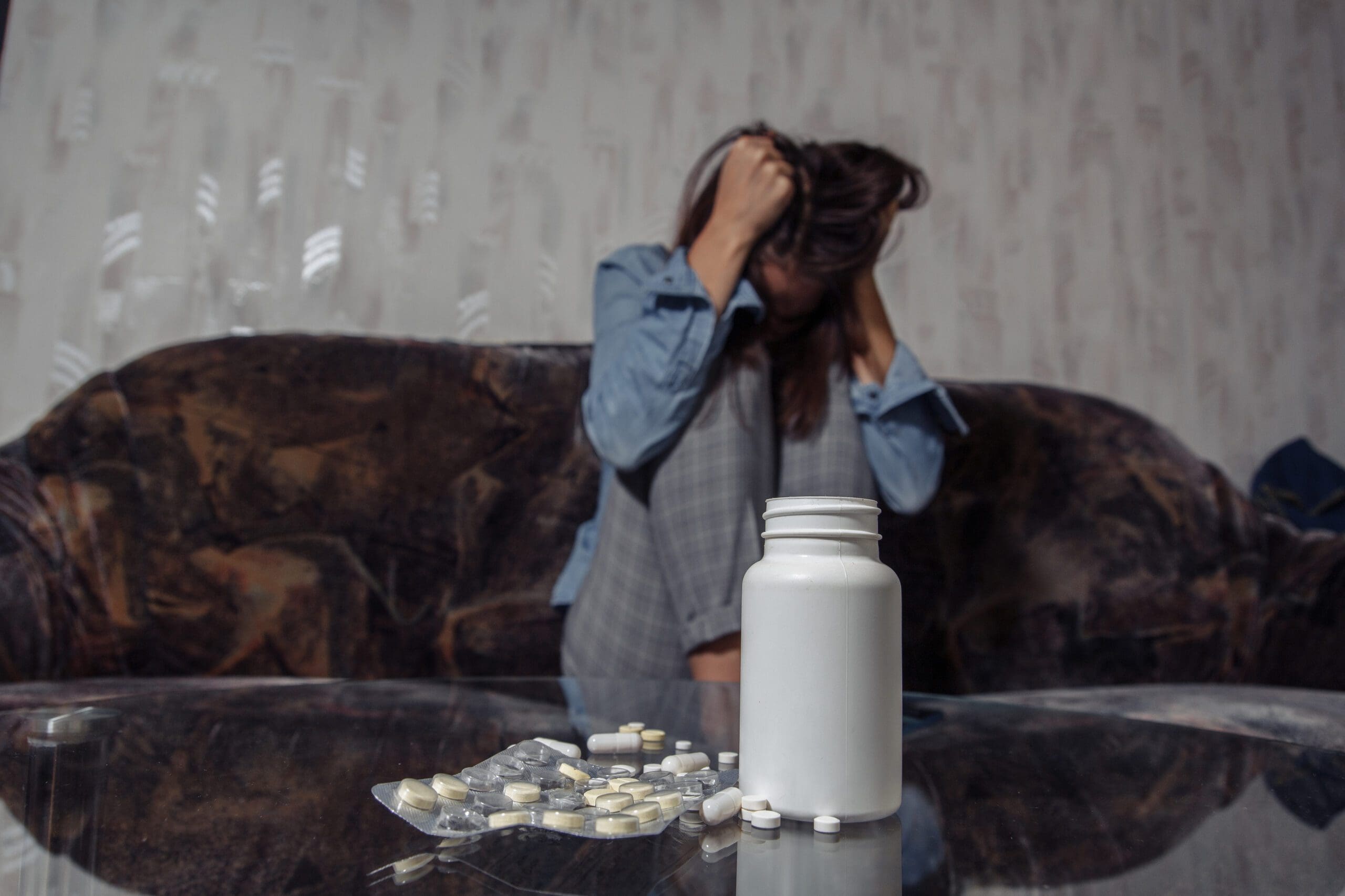 A distressed person sitting behind a table with a spilled bottle of pills in the foreground.