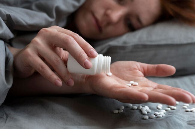 A person is pouring pills from a bottle into their hand while lying in bed.