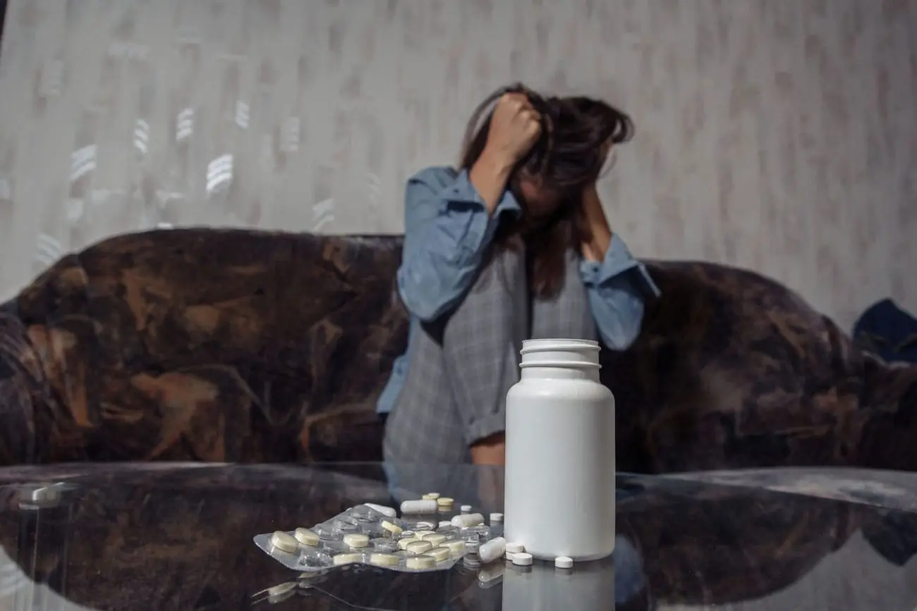 A person with long hair, sitting on a dark sofa, appears distressed, clutching their head with both hands. In the foreground, an open white pill bottle and several scattered pills lie on a glass table—a stark reminder of the struggles many face in prescription addiction rehab in New Jersey.