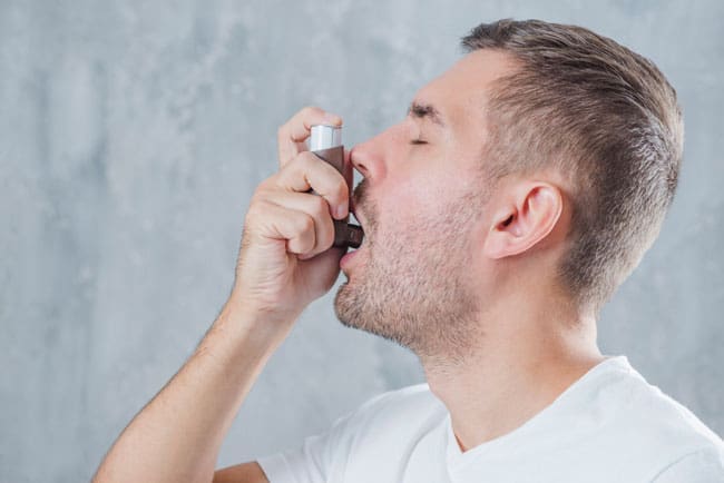A man in a white shirt using an inhaler for asthma or respiratory issues against a textured gray background.