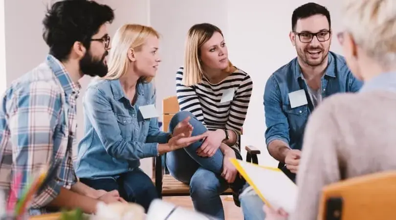 A group of five people sits in a semi-circle, actively engaging in a discussion. Four of them are facing a person holding papers. They appear to be in a casual setting, wearing comfortable clothing with name tags on their shirts, likely part of a drug and alcohol rehab treatment program.