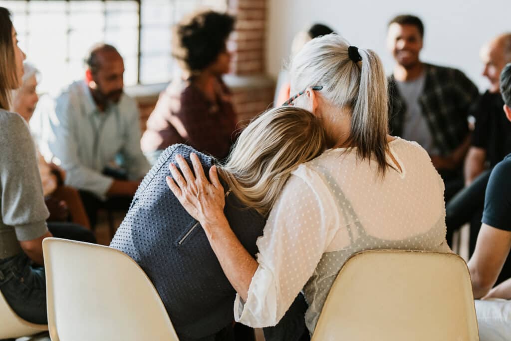 Two people sitting close, one with gray hair comforting the other, who has blonde hair and is leaning on their shoulder. They are part of a larger group seated in a circle at a drug and alcohol rehab treatment center in Denville, engaged in conversation. The scene takes place in a well-lit room with a large window in the background.