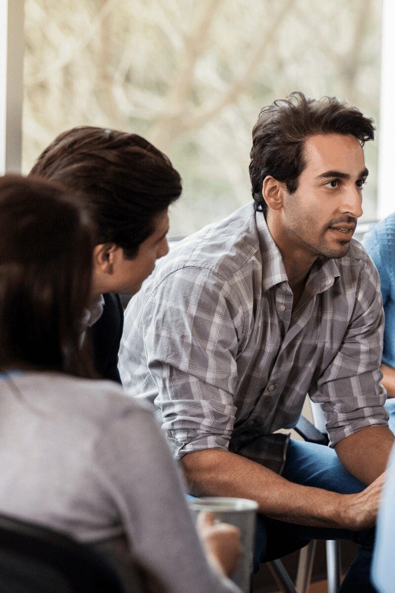 A man with short dark hair, wearing a plaid shirt, is sitting and engaged in conversation with two other people, one of whom is partially visible and wearing a light blue shirt. They seem to be in a casual, indoor setting at a drug and alcohol rehab treatment center in New Jersey, with natural light coming through a window behind them.