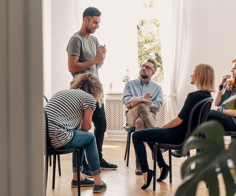 A group of adults in a casual meeting or workshop, one standing and holding a coffee cup, others seated and listening.